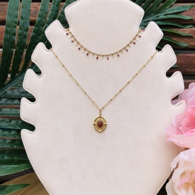 Dois colares dourados num suporte branco com folhas verdes e flores cor de rosa ao fundo