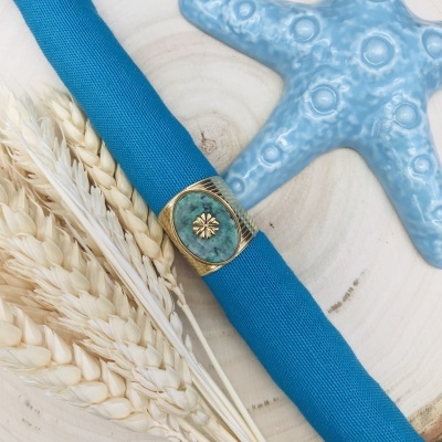 Guardanapo azul com anel decorativo dourado e pedra verde, espigas de trigo e estrela-do-mar azul sobre madeira clara