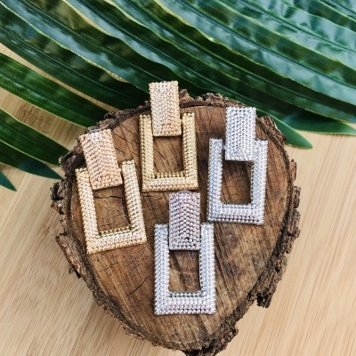 Dois pares de brincos retangulares dourados e prateados sobre madeira com folhas verdes ao fundo