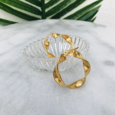 Brincos de ouro texturizados em prato de vidro sobre mármore branco com folhas verdes ao fundo