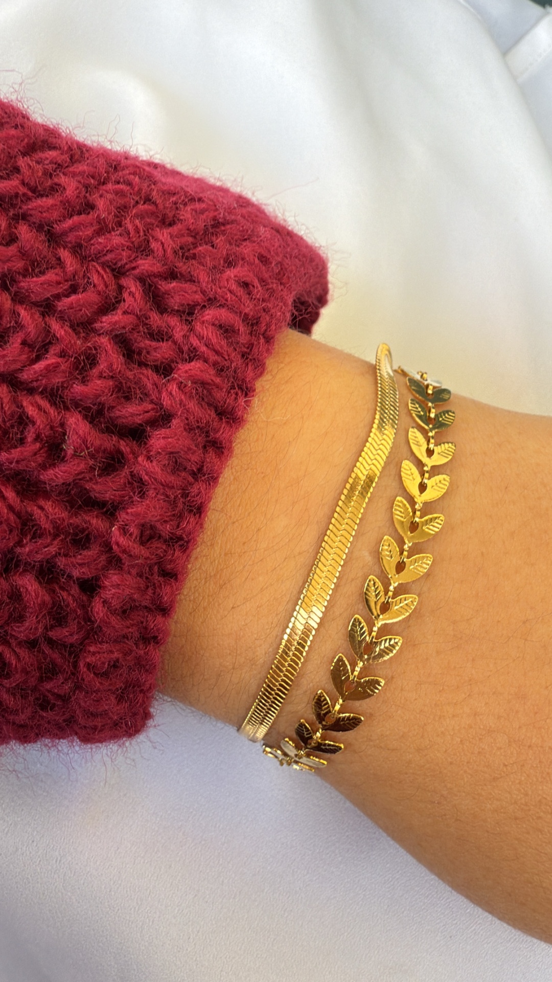 Duas pulseiras douradas com padrões distintos num pulso com manga vinho em malha grossa.