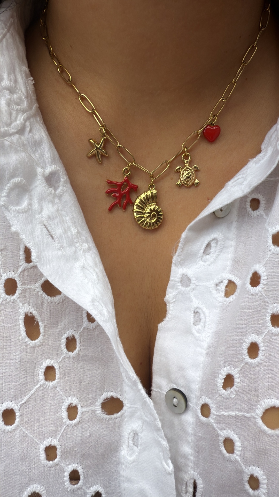 Colar dourado com vários pendentes coloridos e branco, vestido com camisa branca rendada.