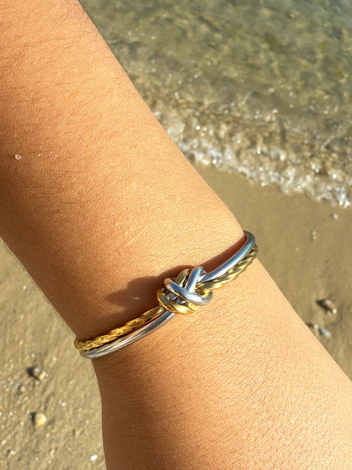 Pulseira de nó dourada e prateada no pulso junto à praia