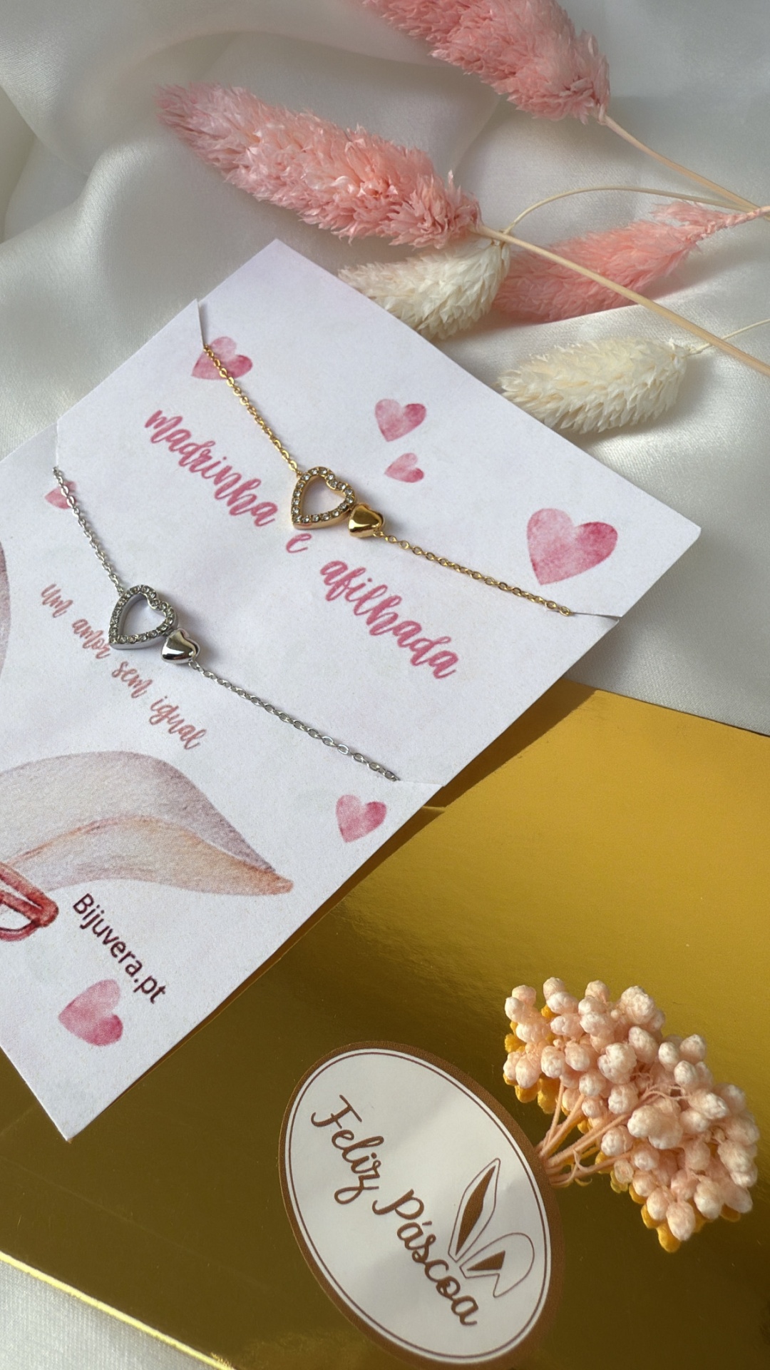 Pulseiras dourada e prateada com corações em cartão sobre tecido branco e caixa dourada com etiqueta Feliz Páscoa