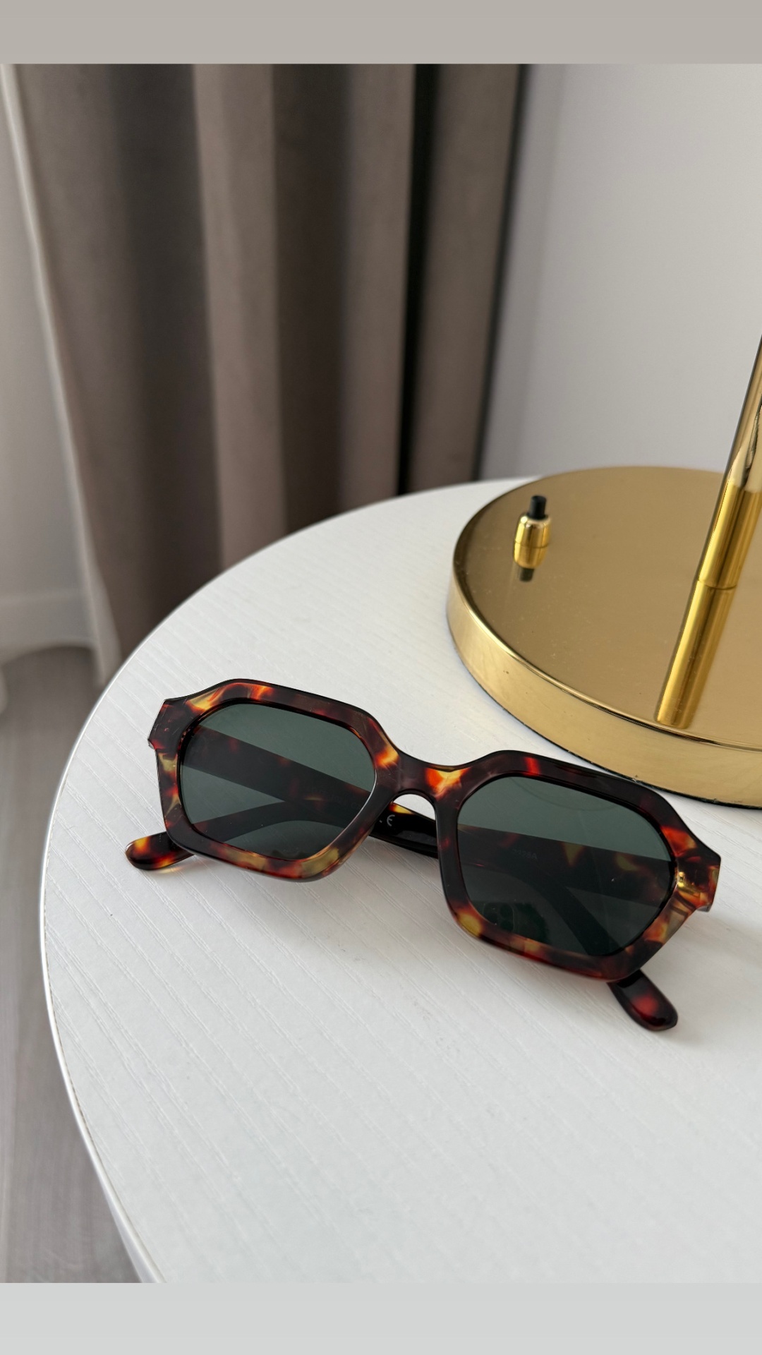 Óculos de sol com armação tartaruga e lentes escuras sobre mesa branca ao lado de candeeiro dourado