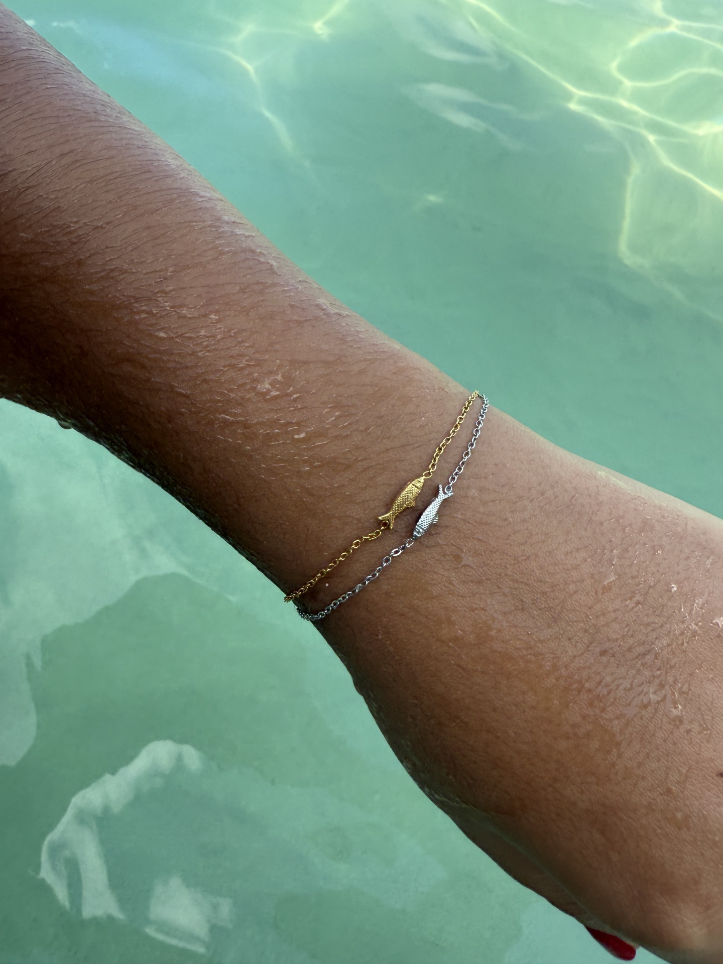 Pulseira dupla dourada e prateada com pássaros no pulso junto a água verde clara