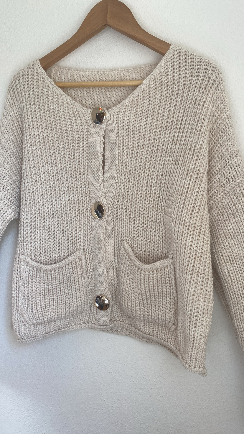 Cardigan bege de malha grossa com botões grandes e bolsos pendurado numa parede branca