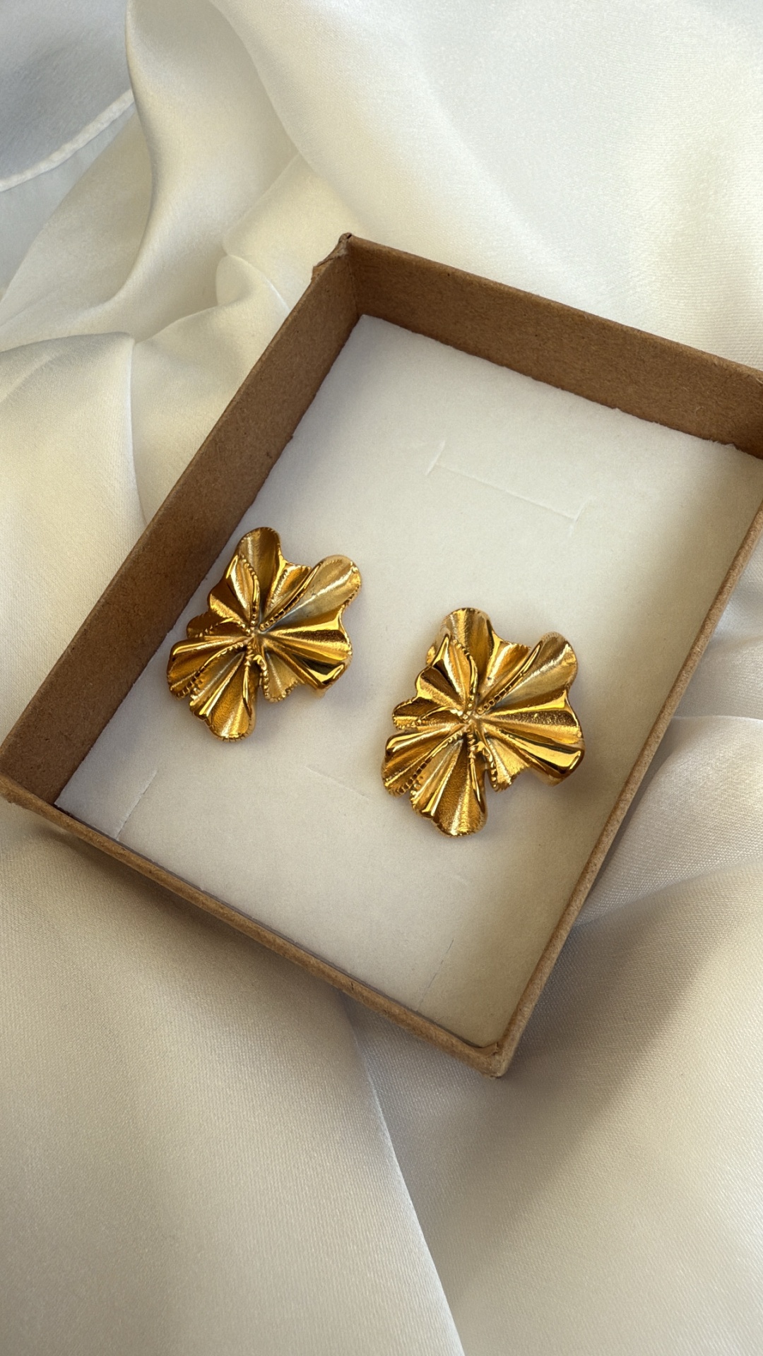 Brincos dourados em forma de folhas numa caixa de cartão sobre tecido de cetim branco