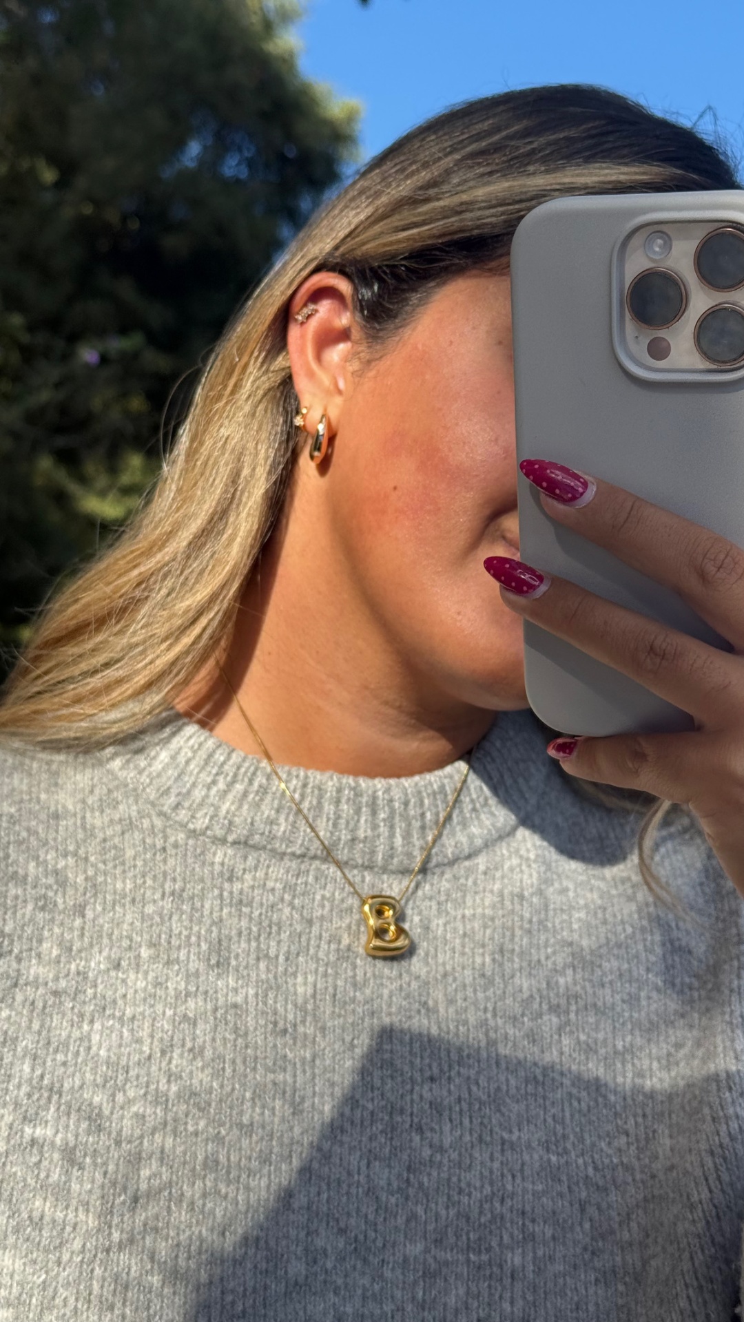 Colar dourado com pendente letra B e brincos dourados em mulher com blusão cinza ao ar livre