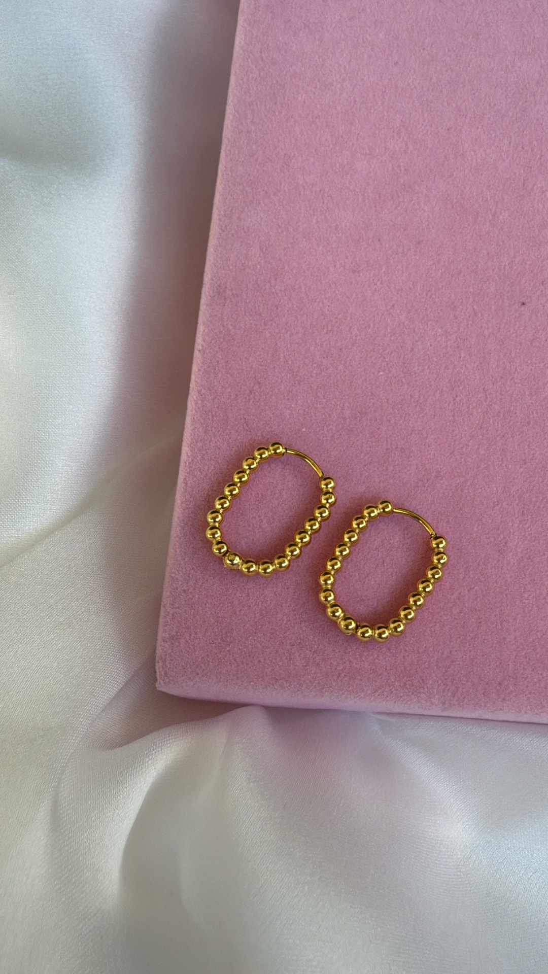 Brincos de argola dourados com contas sobre superfície rosa aveludada e tecido branco