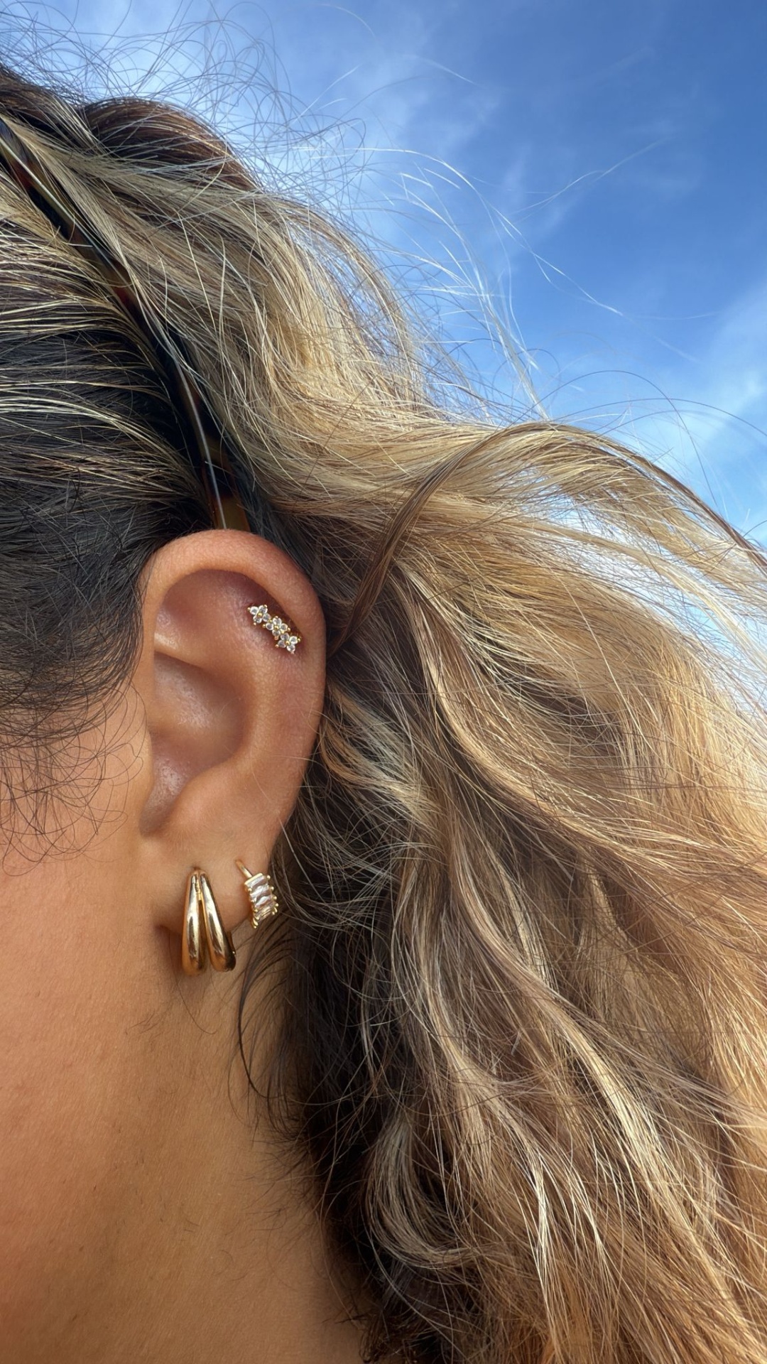Orelha com três brincos dourados e cabelo em fundo de céu azul