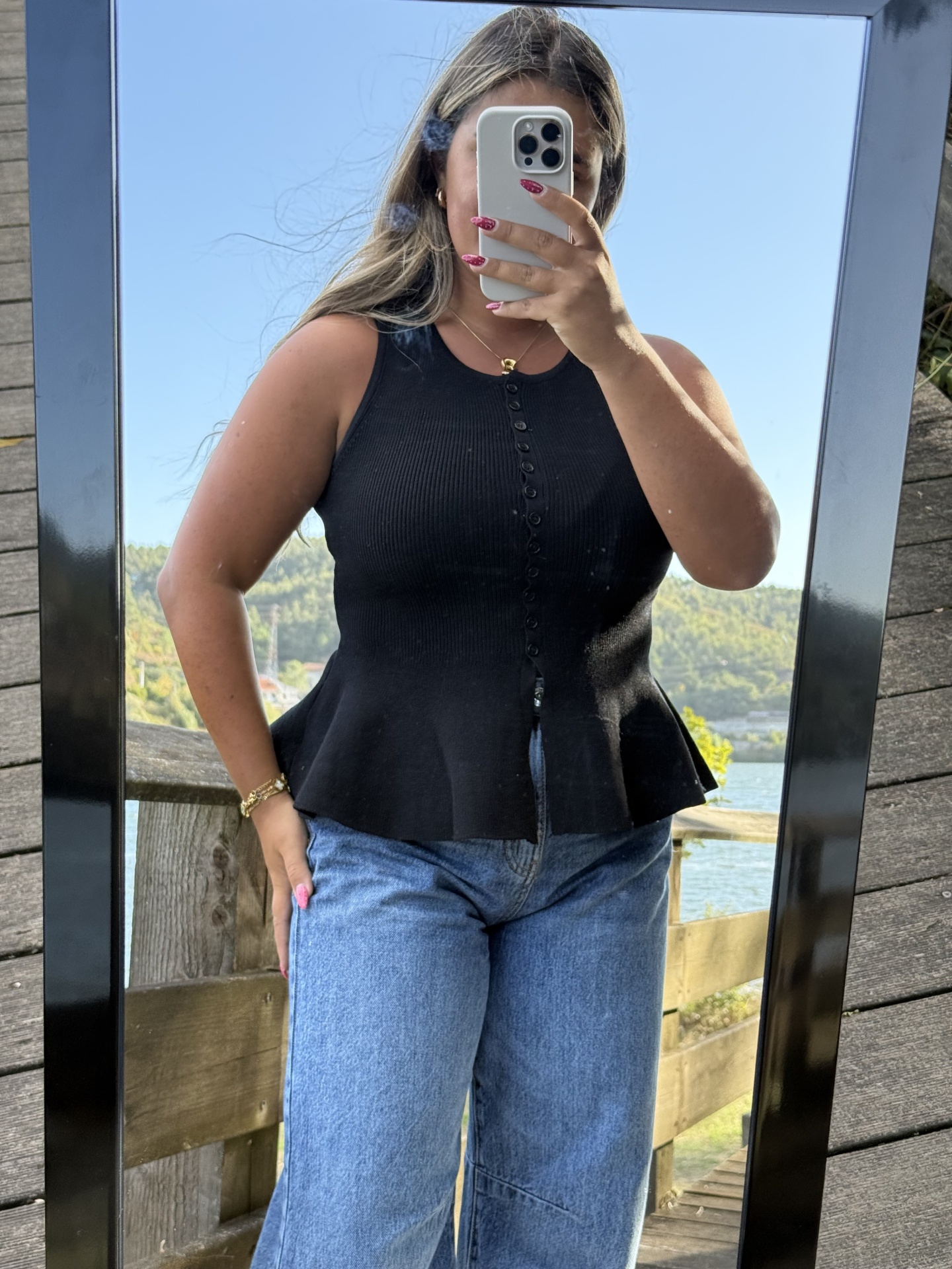 Pessoa a usar top preto de malha com botões e jeans azuis, espelho exterior com paisagem de rio ao fundo
