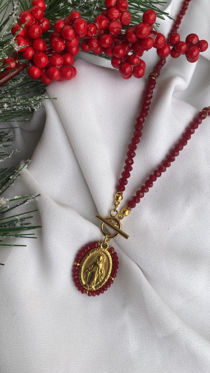 Colar de contas vermelhas com pendente oval dourado em relevo sobre tecido branco com bagas vermelhas e ramos verdes.