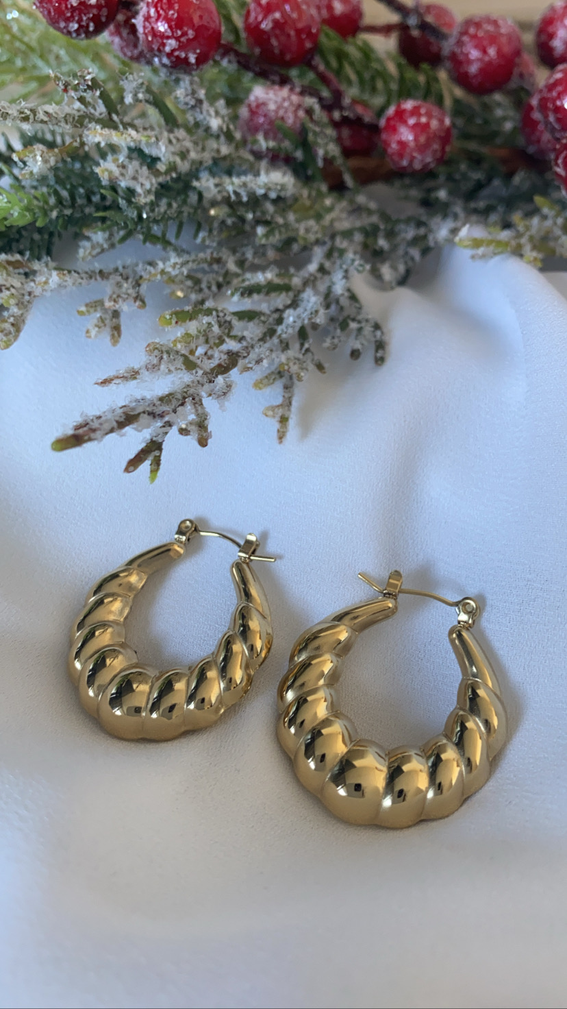 Brincos de argola dourada com textura ondulada sobre tecido branco com decoração de ramos verdes e bagas vermelhas