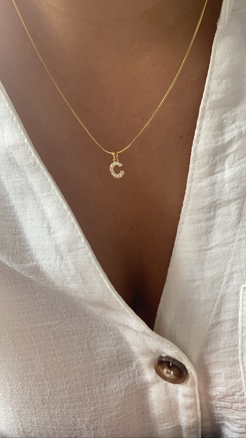 Colar dourado com pendente de letra 'C' em cristais sobre blusa branca
