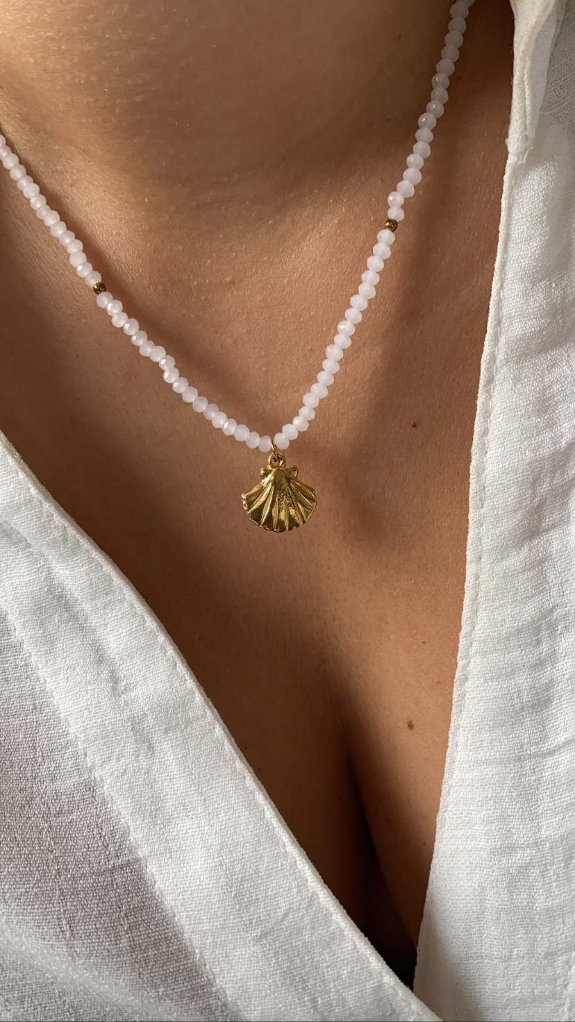 Colar com contas brancas e douradas e pendente dourado em forma de concha sobre blusa branca.