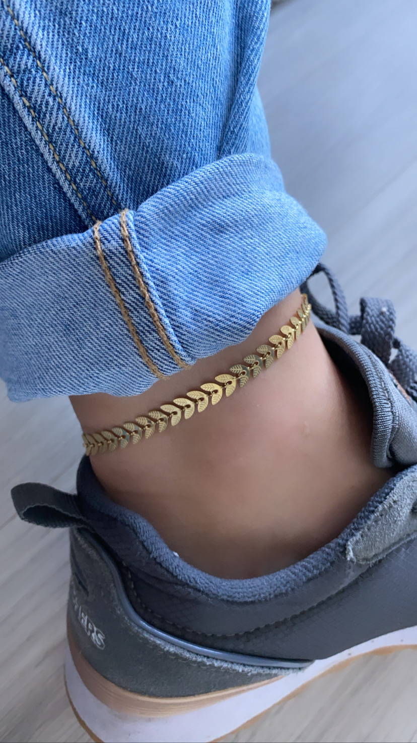 Pulseira de tornozelo dourada com design de folhas usada com calças de ganga e sapatos de desporto cinzentos