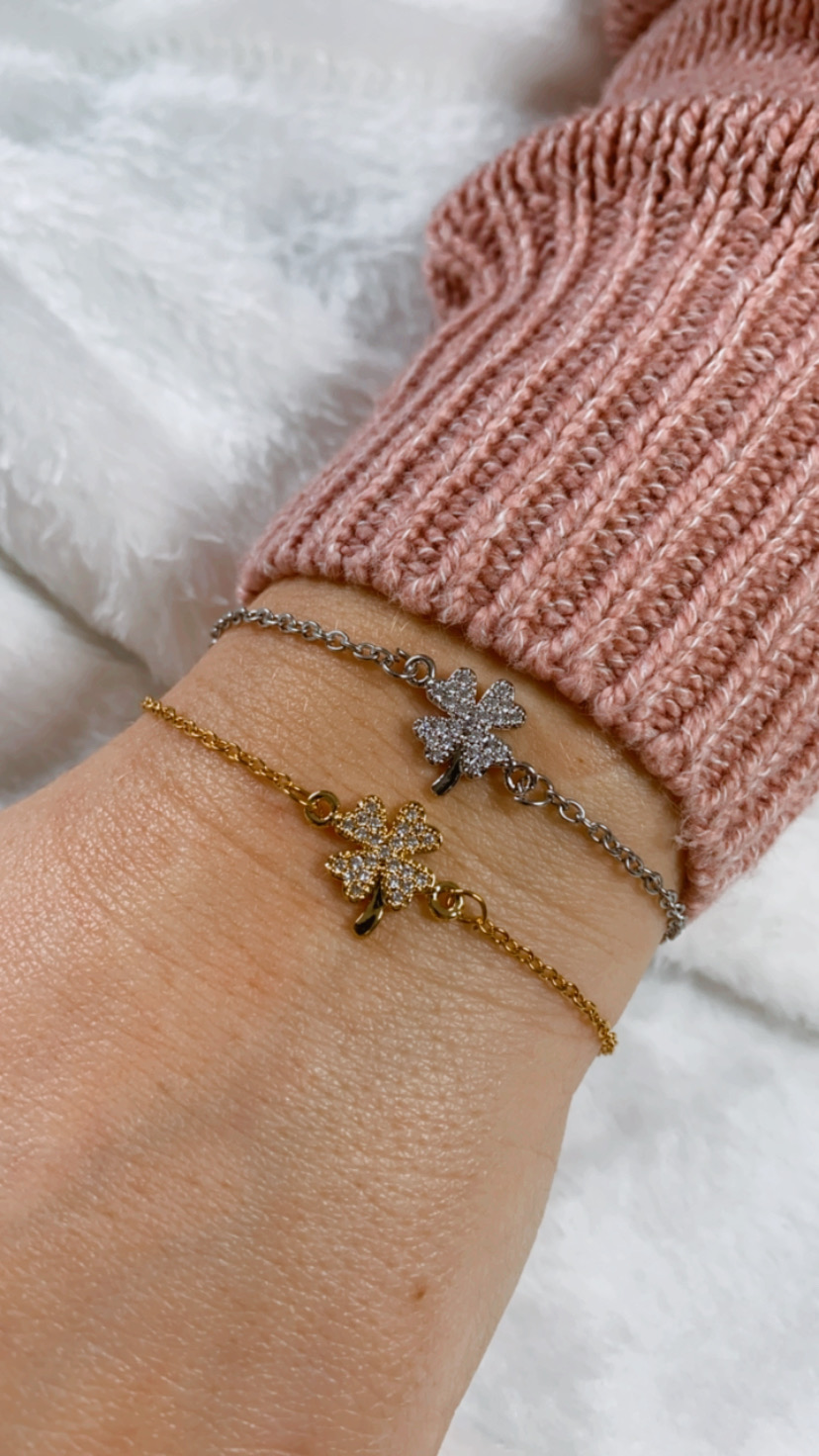 Dois braceletes, dourado e prateado, com pendentes de trevo brilhantes num pulso com camisola rosa