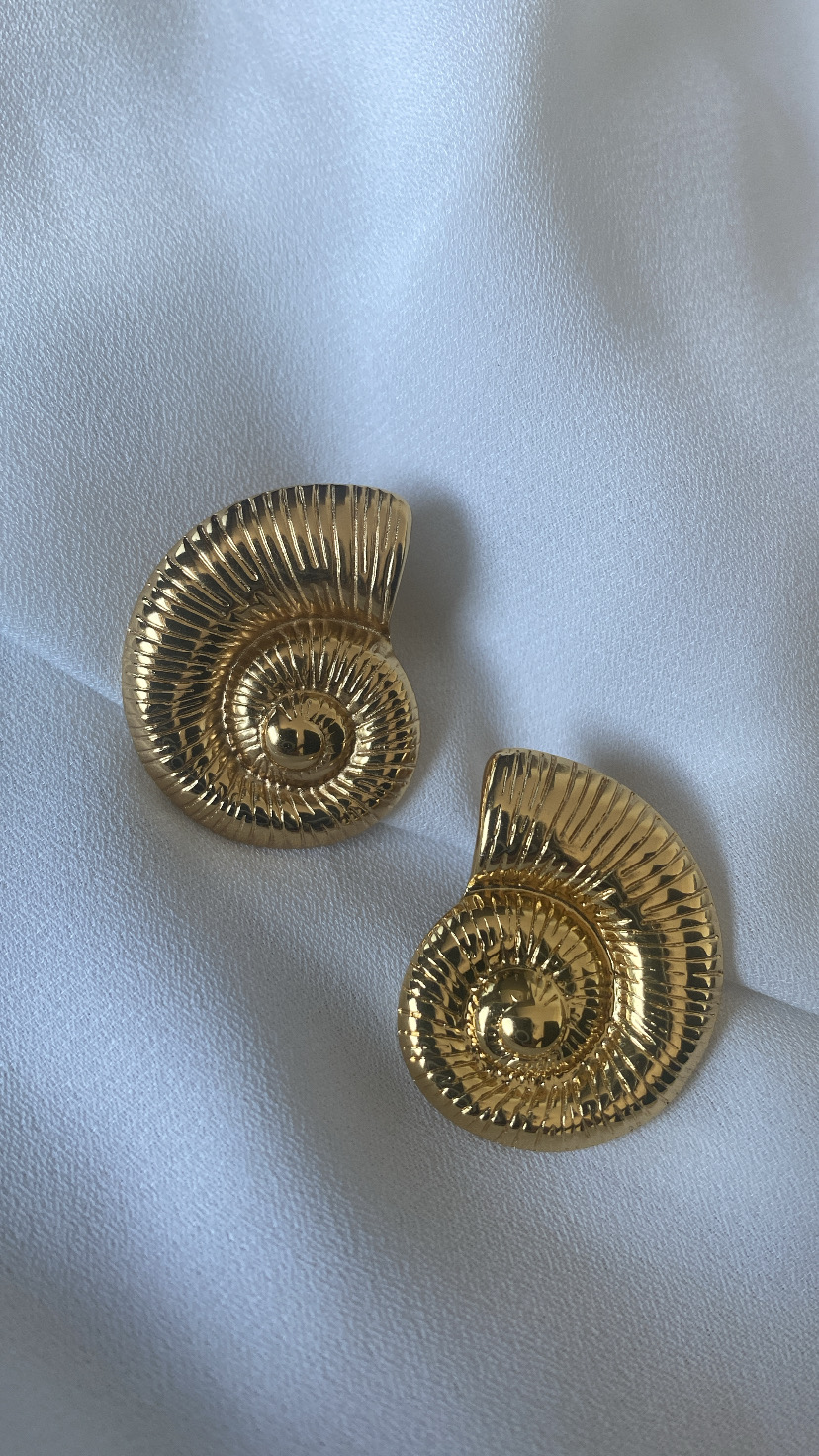 Brincos dourados em forma de concha com textura estriada sobre tecido branco