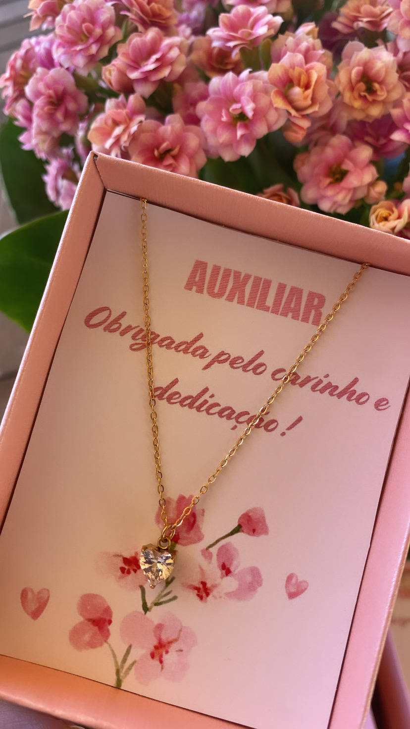 Colar dourado com pendente em pedra transparente em caixa rosa com mensagem de agradecimento e flores ao fundo