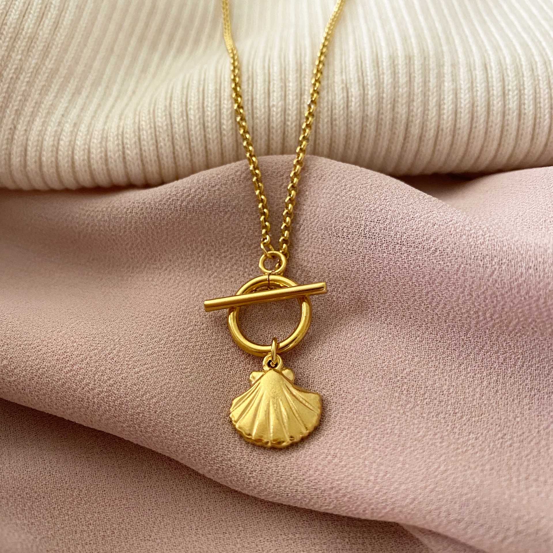 Colar dourado com pendente de concha sobre tecidos creme e rosa.