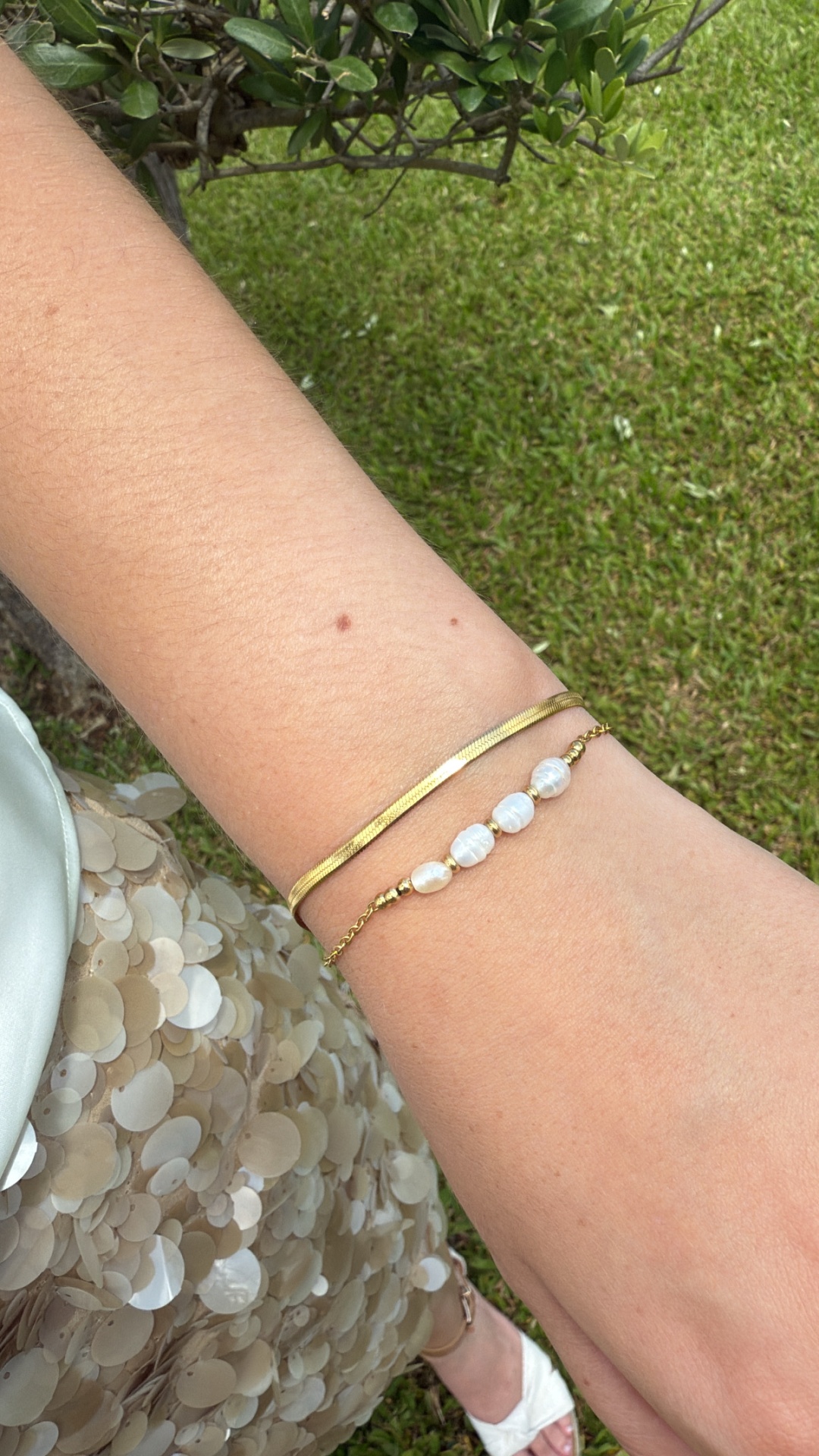 Pulseira dourada lisa e pulseira com contas brancas no pulso num outdoor com relva