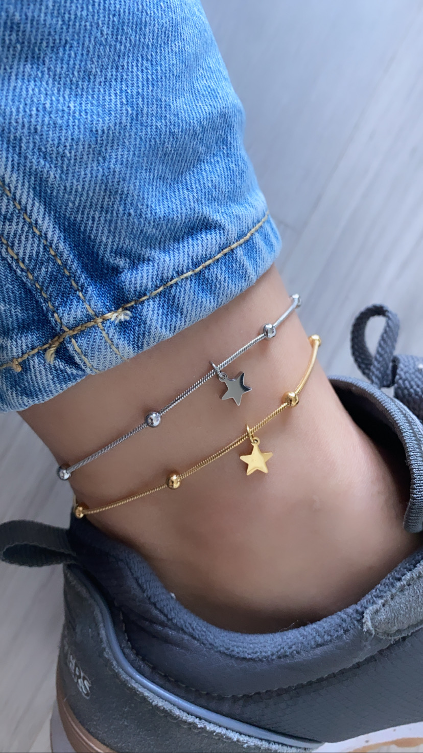 Tornozelo com duas pulseiras finas, uma prateada e outra dourada com pendentes de estrelas, calça de ganga azul e sapatilha cinzenta