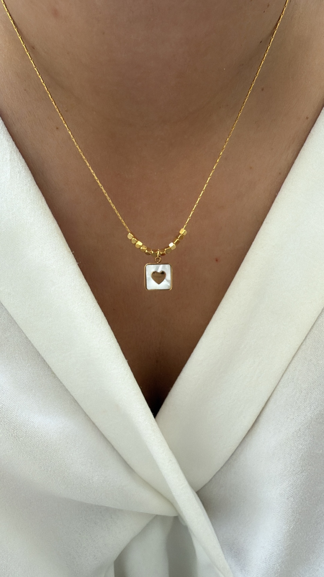 Colar dourado com pingente quadrado e coração recortado numa blusa branca