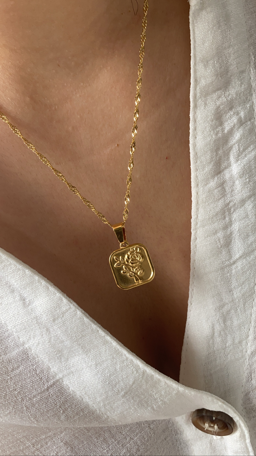 Colar dourado com pendente quadrado de rosa em relevo, sobre camisa branca.