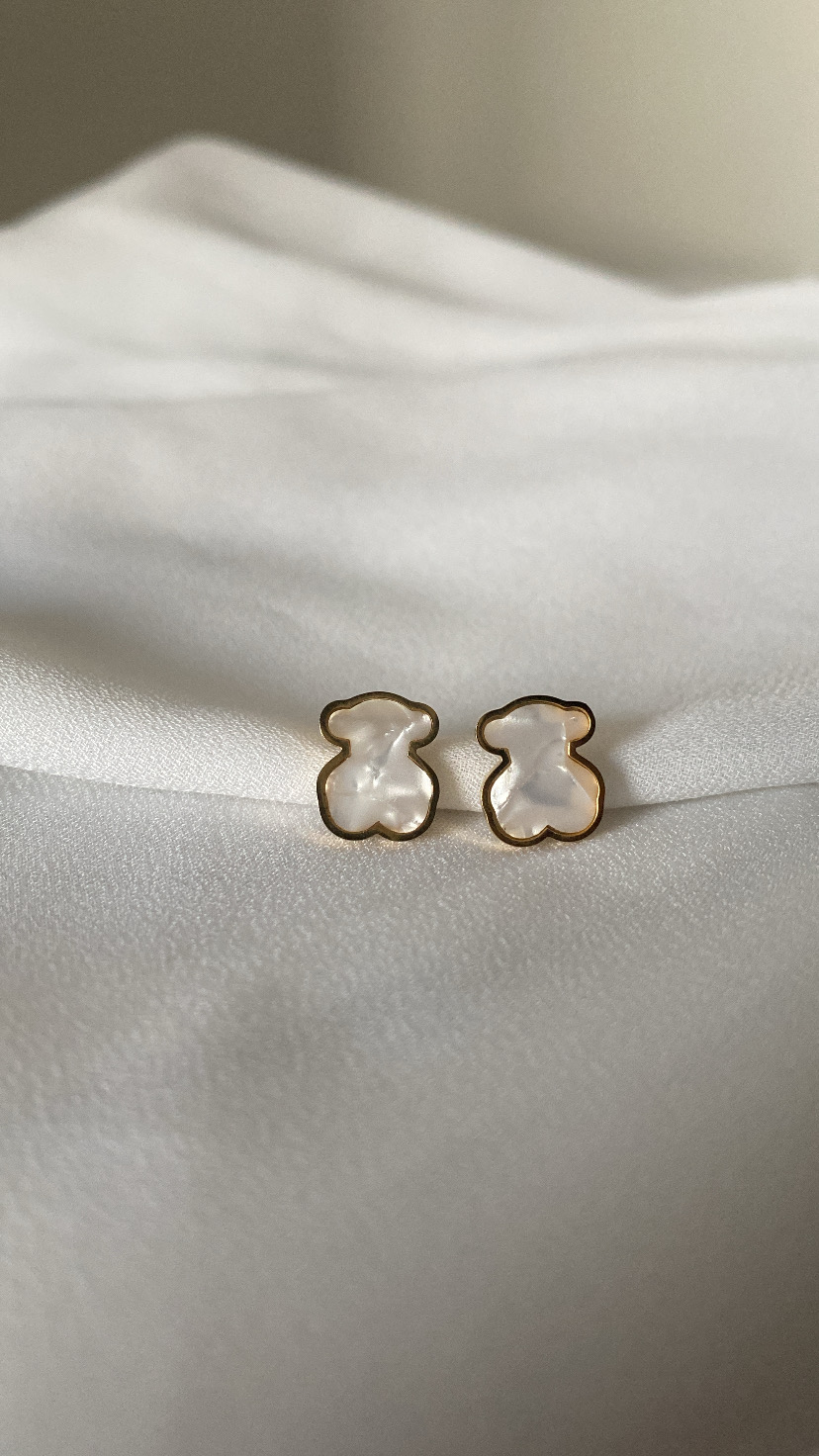 Brincos em forma de ursinho em madrepérola com contorno dourado sobre tecido branco.