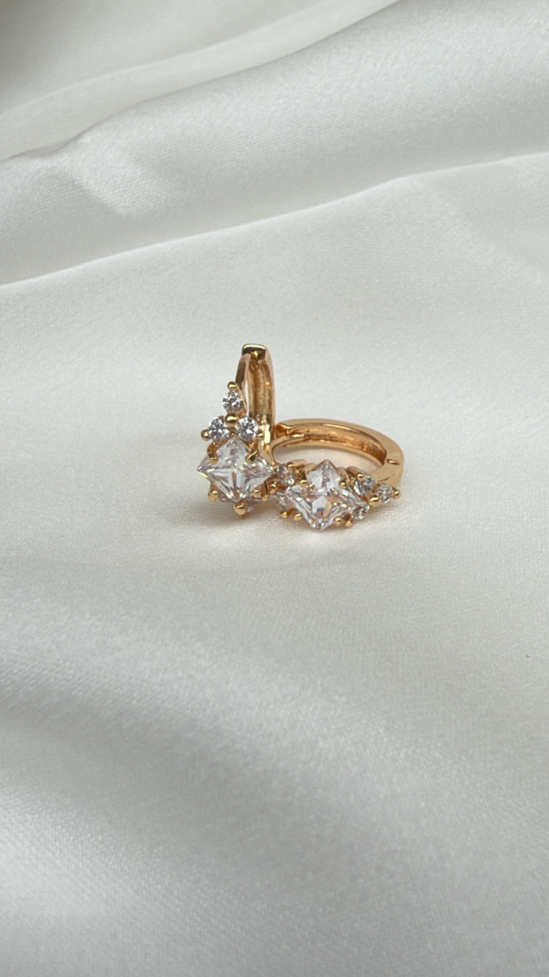 Brincos dourados com pedras quadradas e cristais sobre tecido branco acetinado