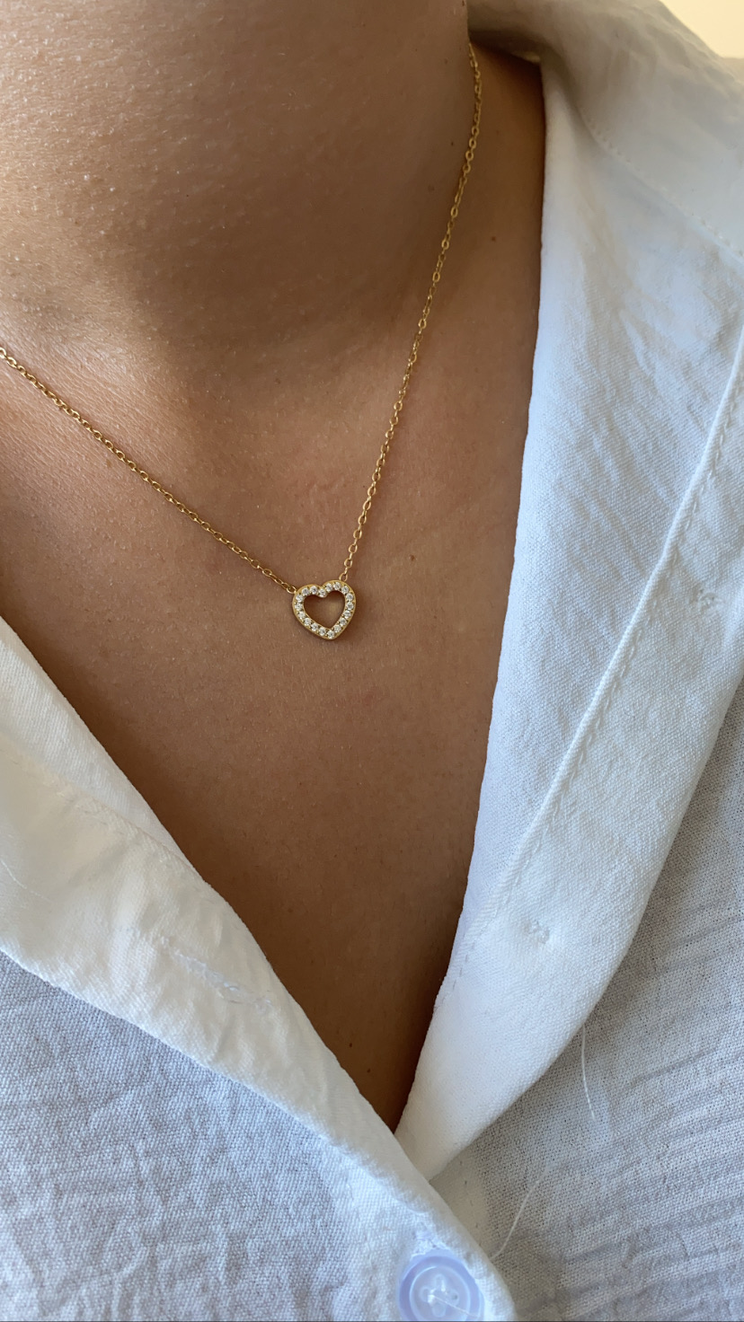 Colar dourado com pendente de coração com cristais sobre camisa branca.