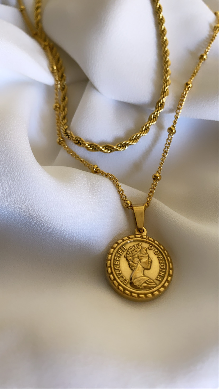 Colar dourado com pendente moeda de Elizabeth II sobre tecido branco.