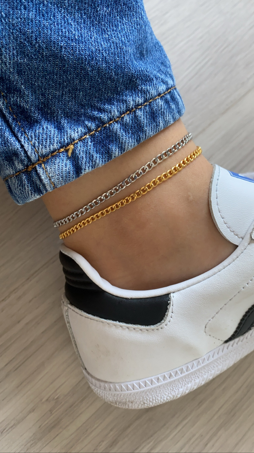 Tornozelo com pulseiras prateada e dourada, jeans azul e ténis brancos com detalhes pretos.