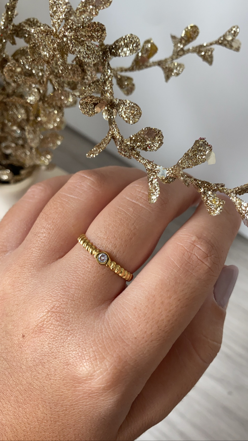 Anel dourado com pedra pequena numa mão perto de decoração dourada.