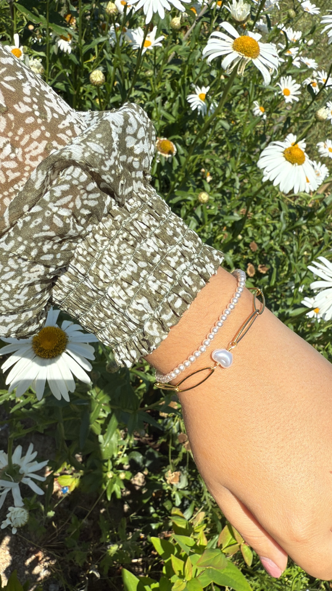 Pulseira com contas de pérolas e bracelete dourada com pingente de concha num pulso junto a flores margueritas