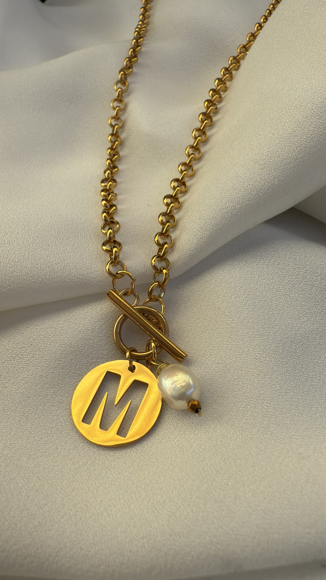 Colar dourado com pendente letra M e pérola sobre tecido creme.