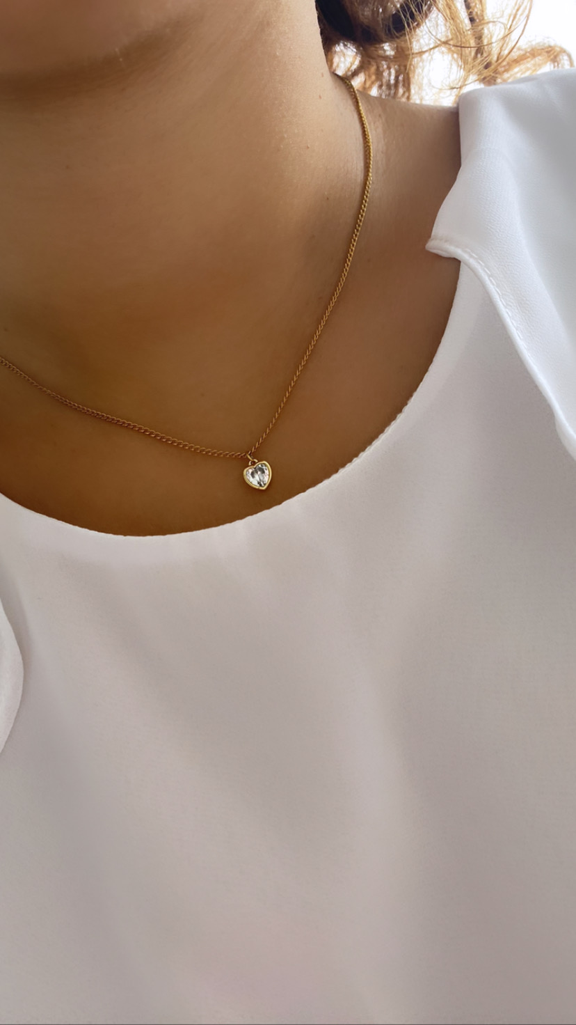 Colar dourado com pendente em forma de coração transparente numa pessoa com blusa branca