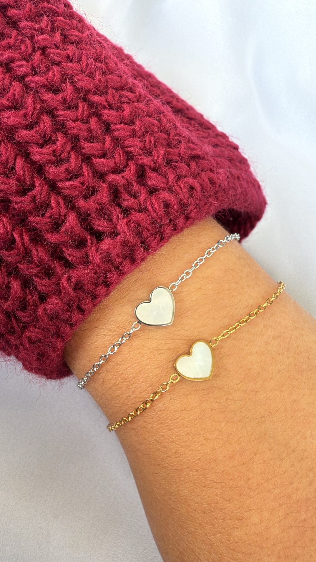 Duas pulseiras delicadas, prata e dourado, com corações brancos, no pulso de uma pessoa com camisola vermelha.