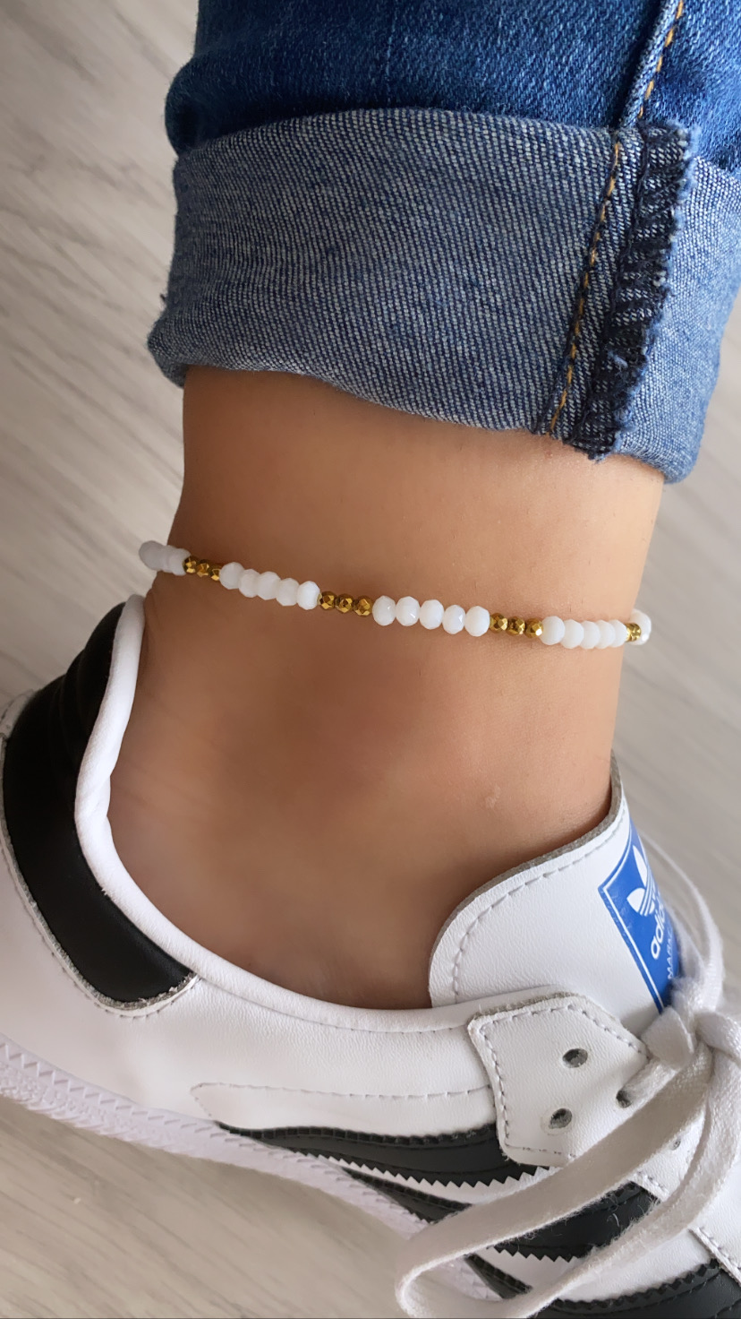 Tornozeleira com contas brancas e douradas no tornozelo com calças de ganga e ténis branco e preto