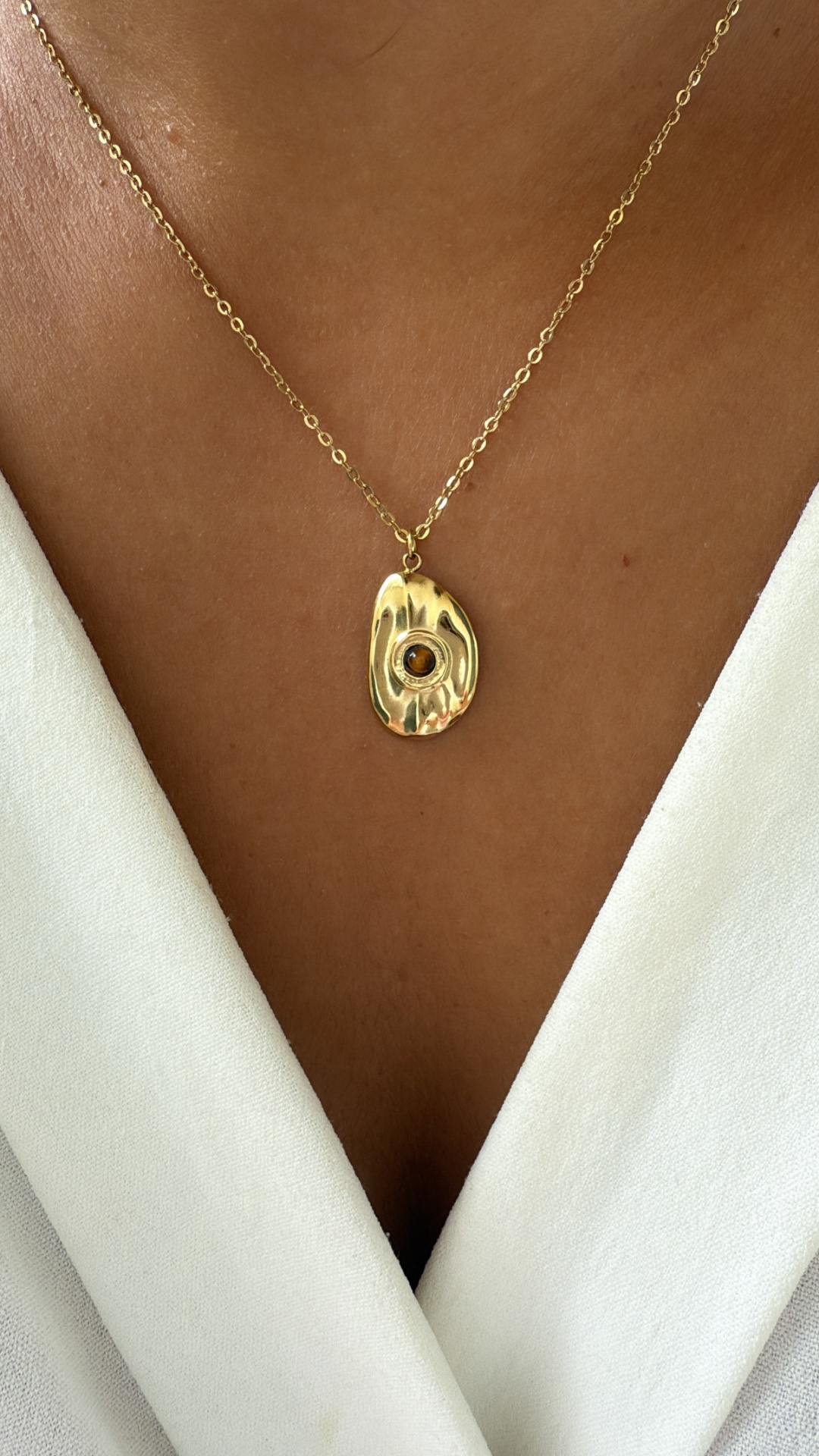 Colar dourado com pendente texturizado e pedra âmbar, usado com camisa branca.