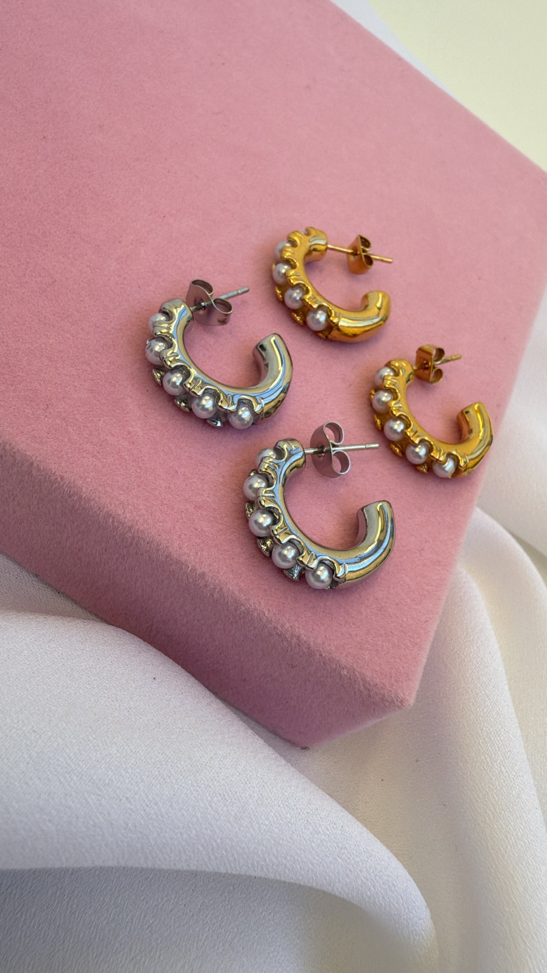 Dois pares de brincos meia argola com pérolas, um prateado e outro dourado sobre veludo rosa