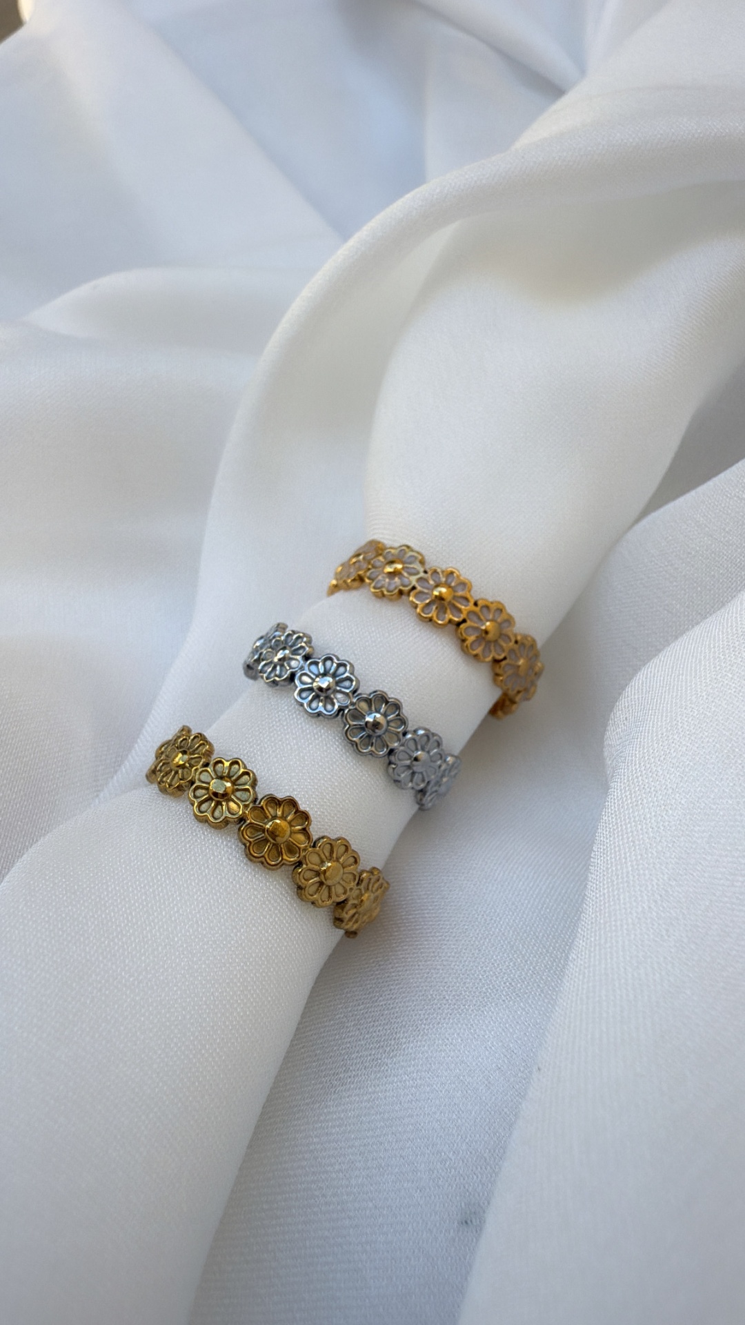 Três anéis florais metálicos em dourado e prateado sobre tecido branco.