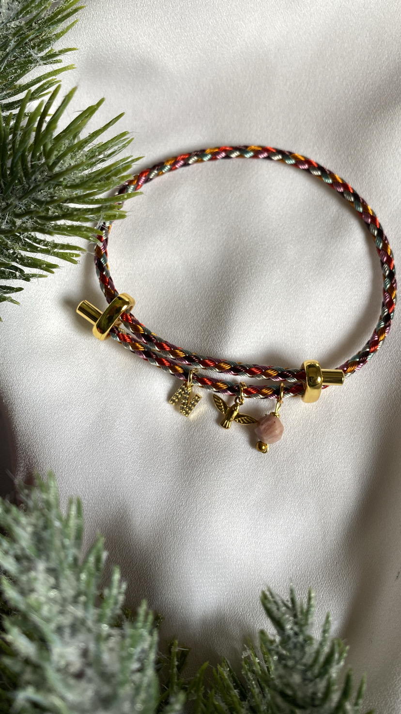 Pulseira trançada colorida com pingentes dourados e pedra rosa sobre tecido branco