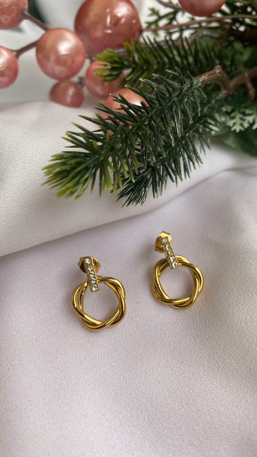 Brincos dourados entrelaçados com cristais sobre tecido branco e decoração verde e rosa