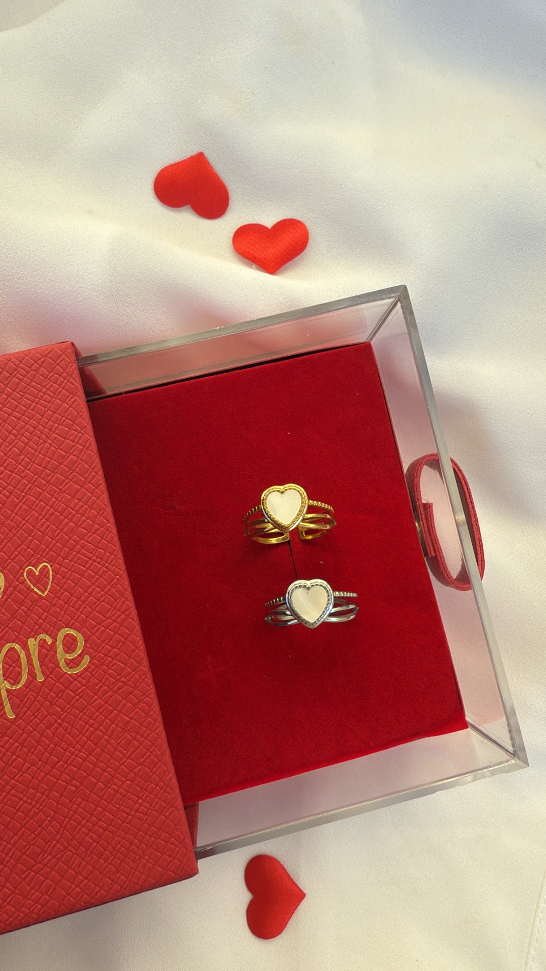 Dois anéis em forma de coração dentro de caixa transparente com base vermelha, caixa vermelha com a palavra Amore em dourado e corações vermelhos decorativos sobre tecido branco.