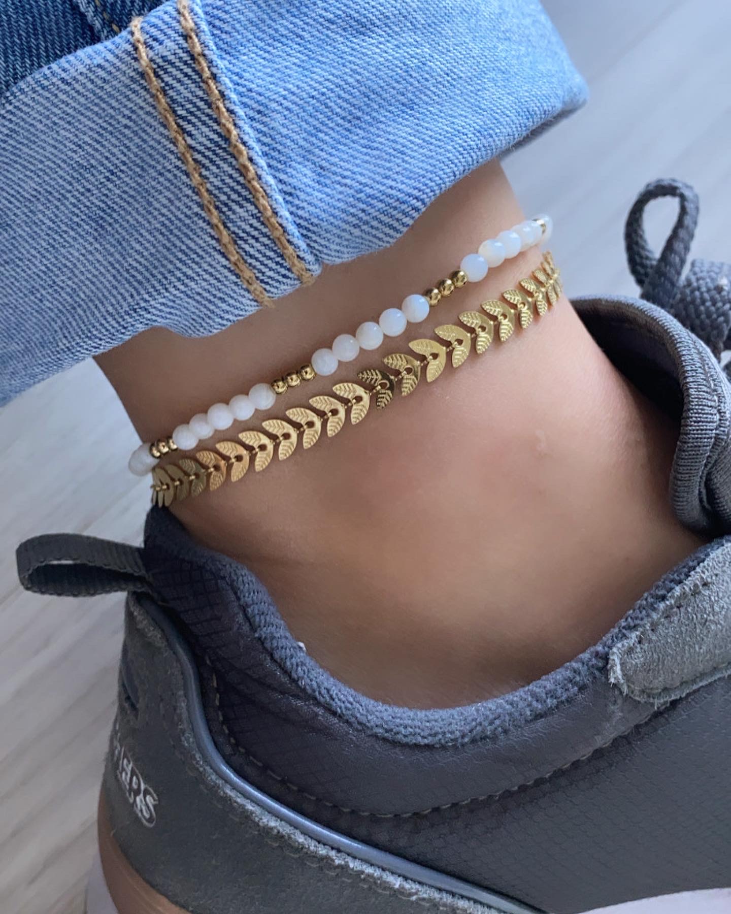 Tornozelo com duas pulseiras douradas e brancas, calçado desportivo cinzento e calças de ganga.