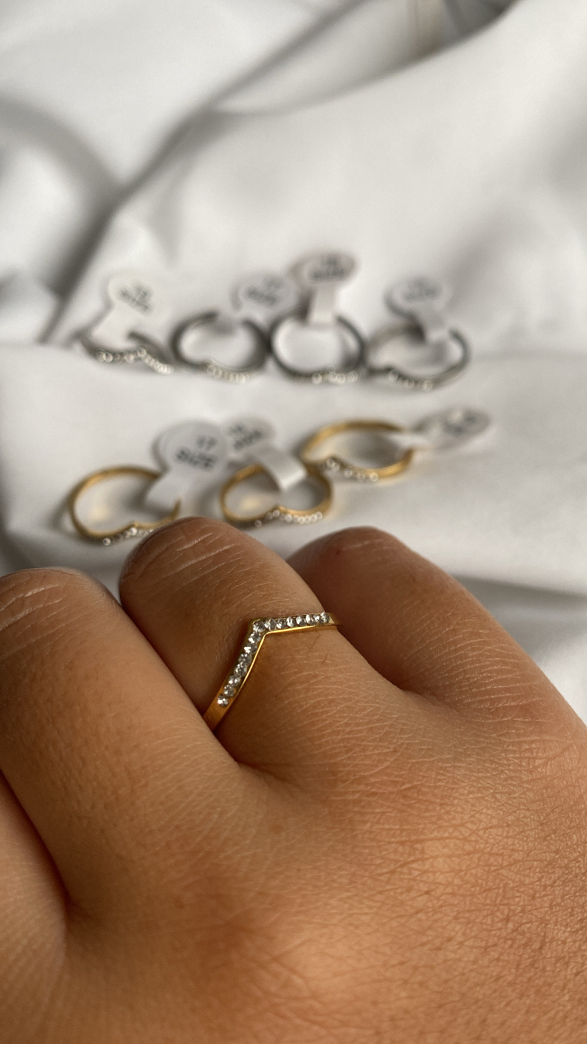 Anel dourado com pedras brilhantes em formato V na mão, outros anéis semelhantes ao fundo