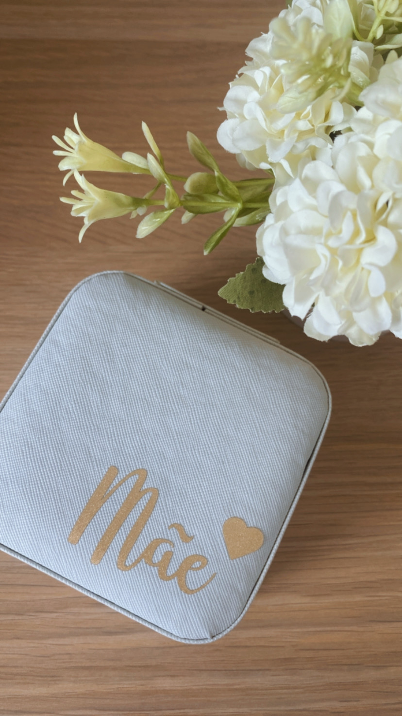 Estojo cinza claro com texto dourado 'Mãe' ao lado de flores brancas