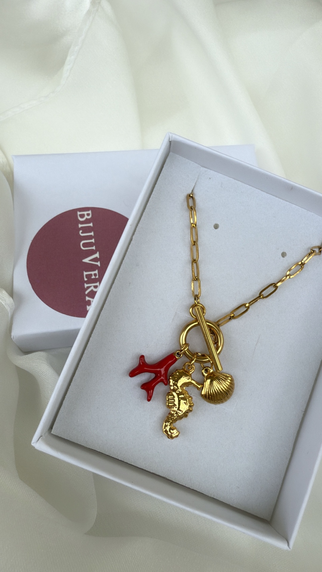 Colar dourado com pendentes de cavalo-marinho, concha e coral numa caixa branca com etiqueta BIJUVERA.